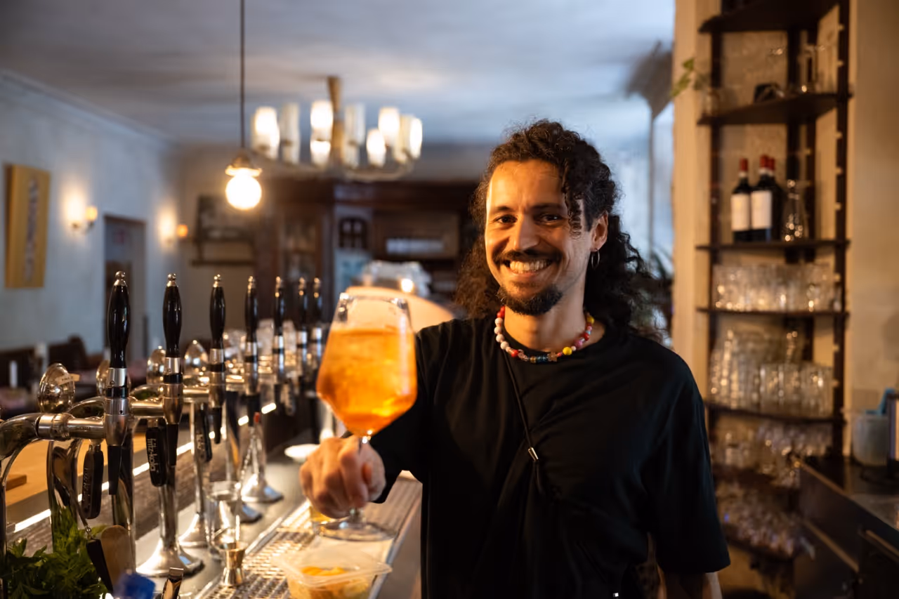 Mann mit schwarzem T-Shirt und Perlenkette hält ein Glas mit orangefarbenem Getränk an der Bar.
