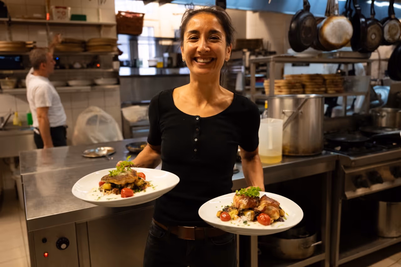 Lächelnde Kellnerin trägt zwei Teller mit angerichtetem Essen in einer Restaurantküche.