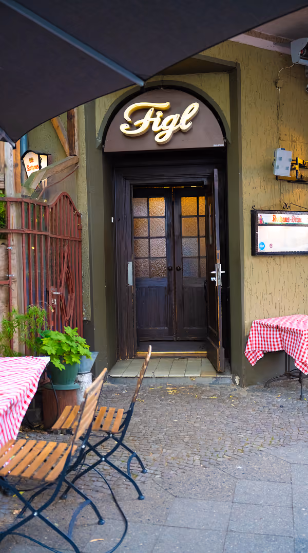 Entrance to Figl restaurant with dark wooden double doors, outdoor tables with red and white checkered tablecloths, and an open umbrella.