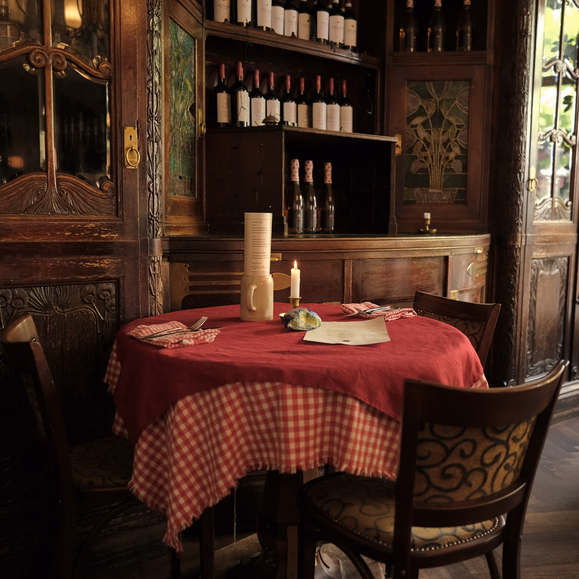 Gemütlicher Tisch mit rotem und kariertem Tischtuch, Kerze, Menü und Weinflaschen im Holzregal.