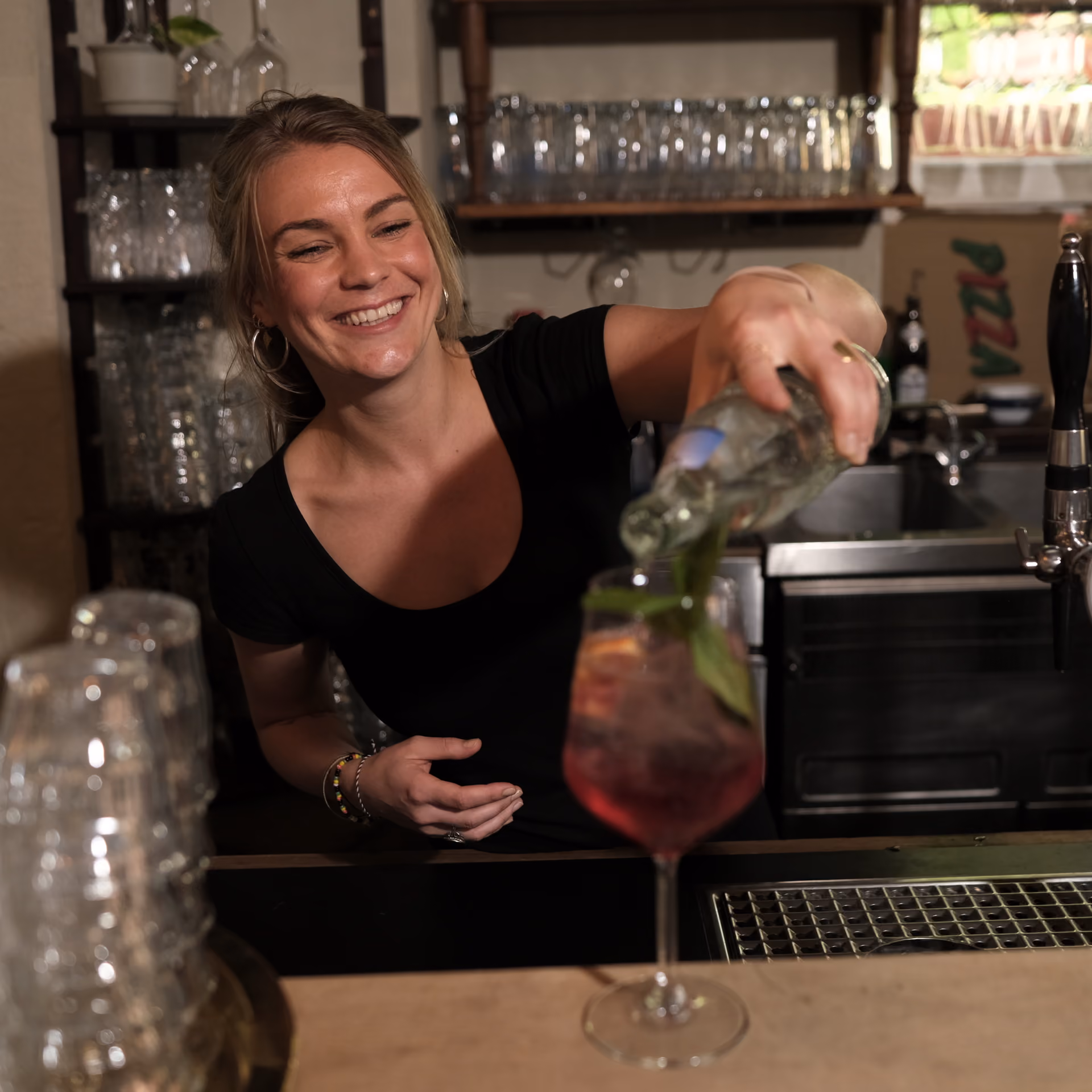 Lächelnde Kellnerin gießt ein Getränk mit frischen Kräutern in ein Glas am Tresen.