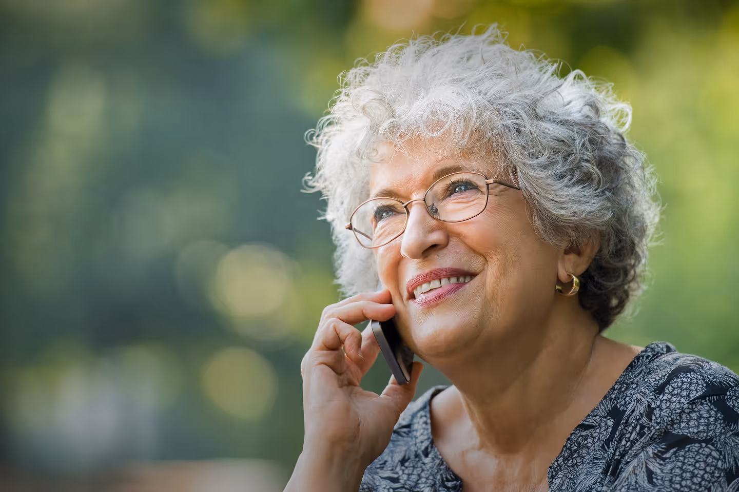Smiling older woman with gray curly hair speaking on smartphone outdoors, wearing glasses – staying in touch with family