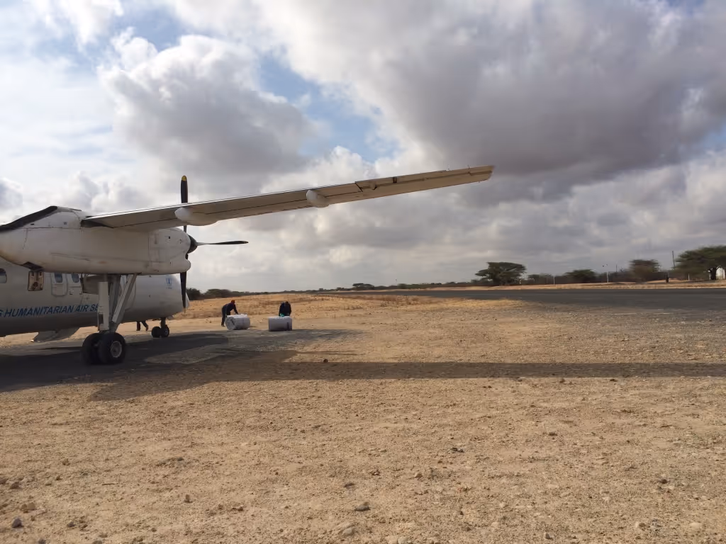 A plane sitting on a runway in an arid environment. Two workers are seen rolling barrels of fuel toward the plane.