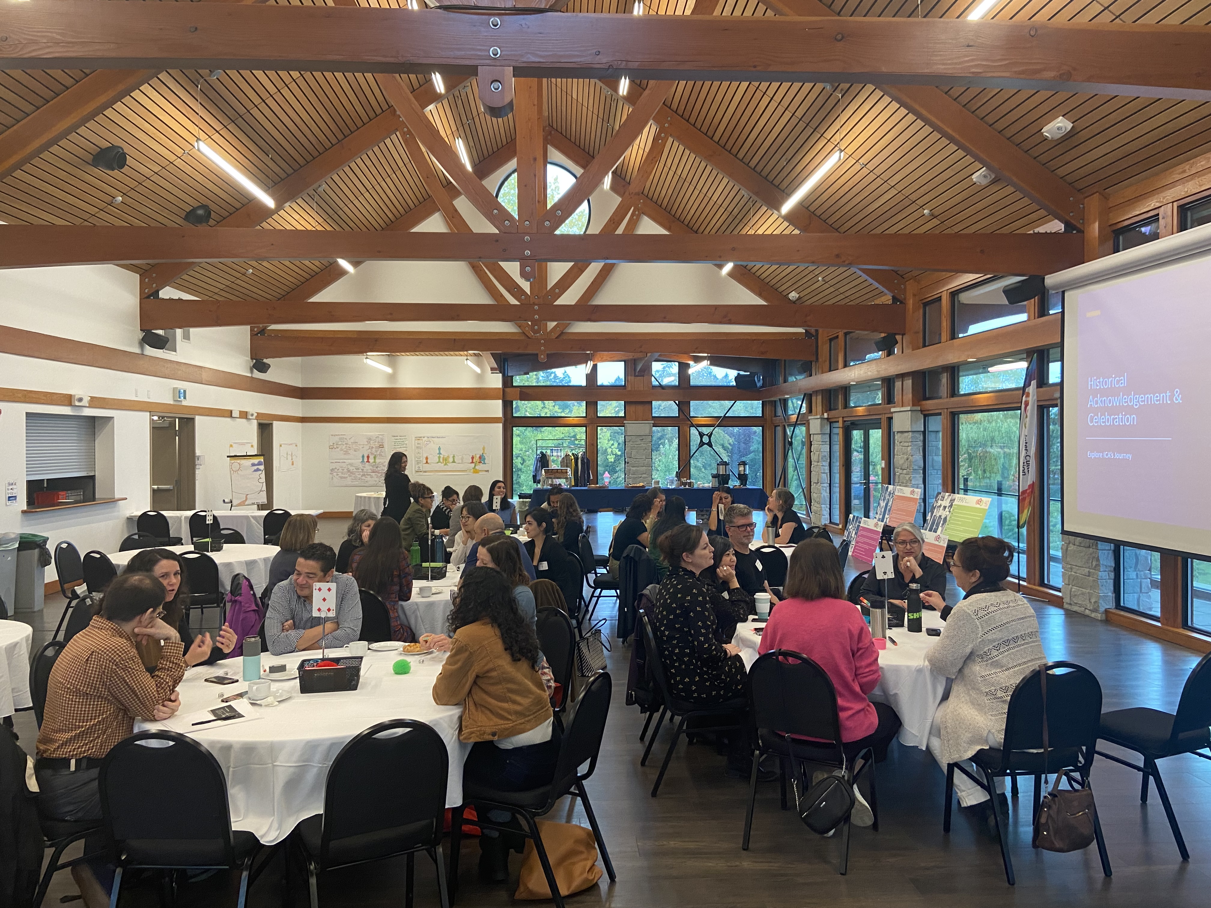A timber frame conference room is filled with round tables and chairs. There are people around each table chatting with each other.
