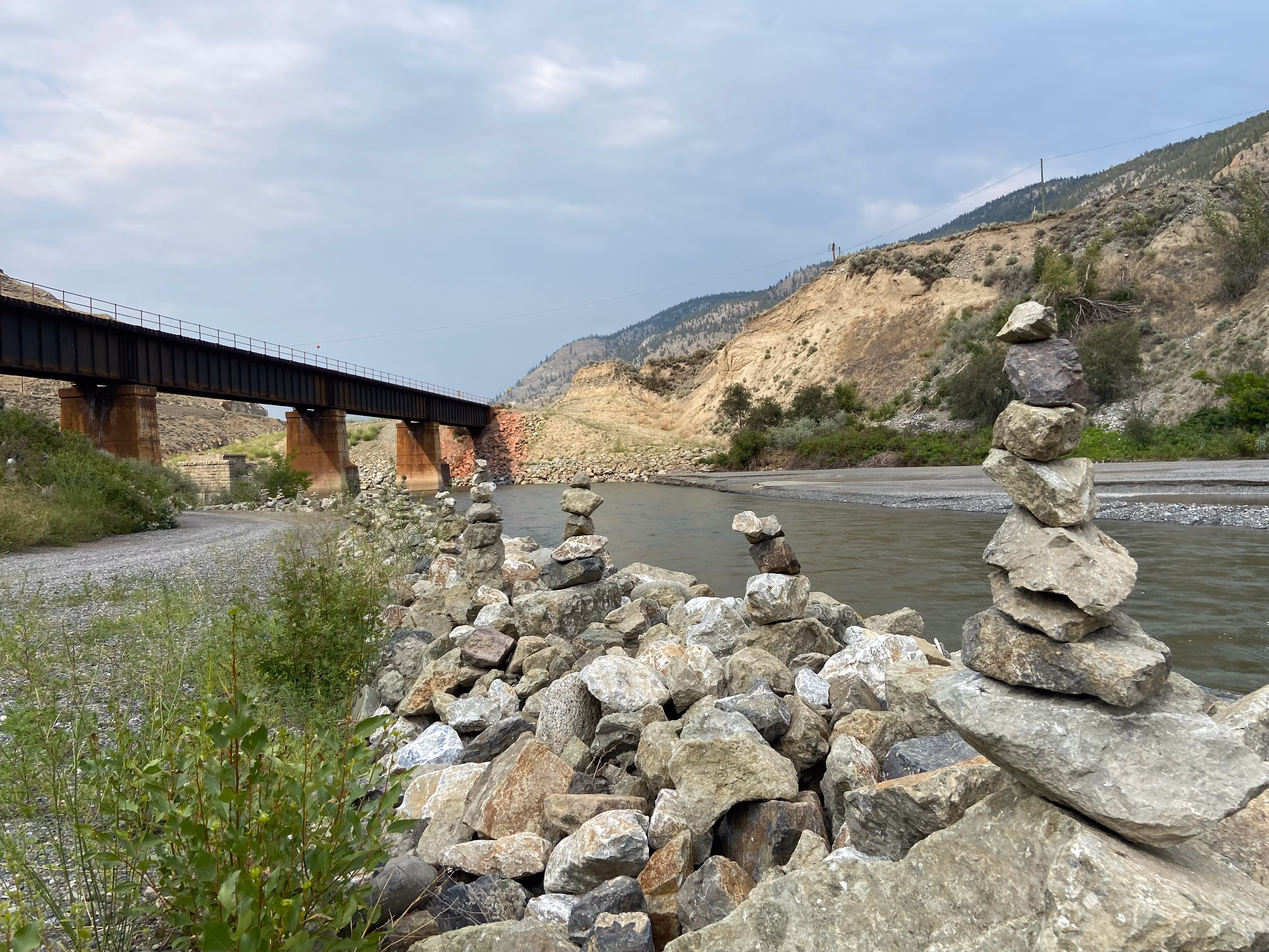 Several stacks of small rocks are seen lining a river. Hills and a big steel rail bridge are seen in the background.