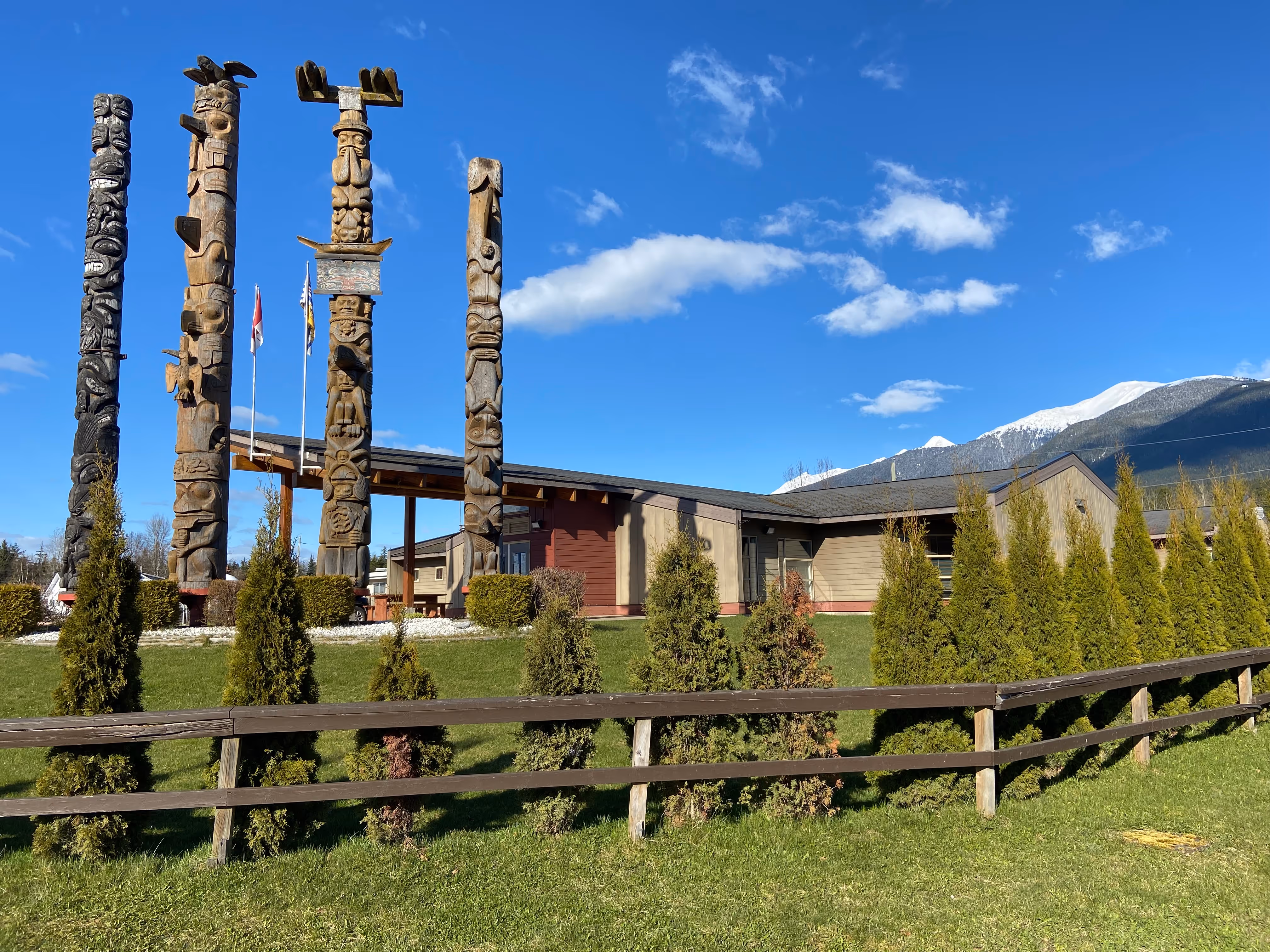 Several wood totem polls tower above a small administrative building. The sky is blue and the grass below is a rich green. 