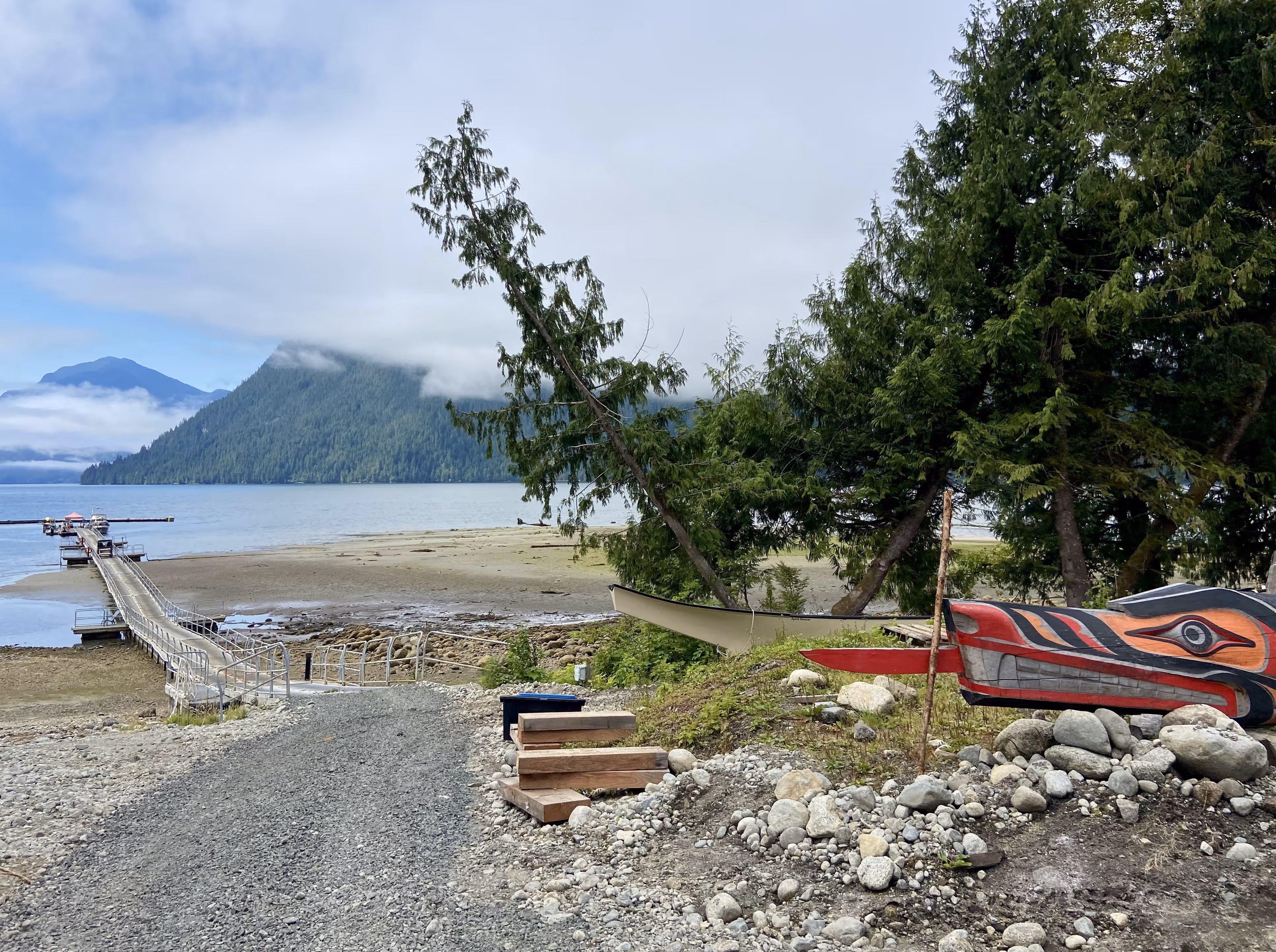 A gravel path leads to a long dock that sits in an ocean inlet in the pacific northwest. In the foreground is a wood sculpture of indigenous style and in the background are tree covered mountains.