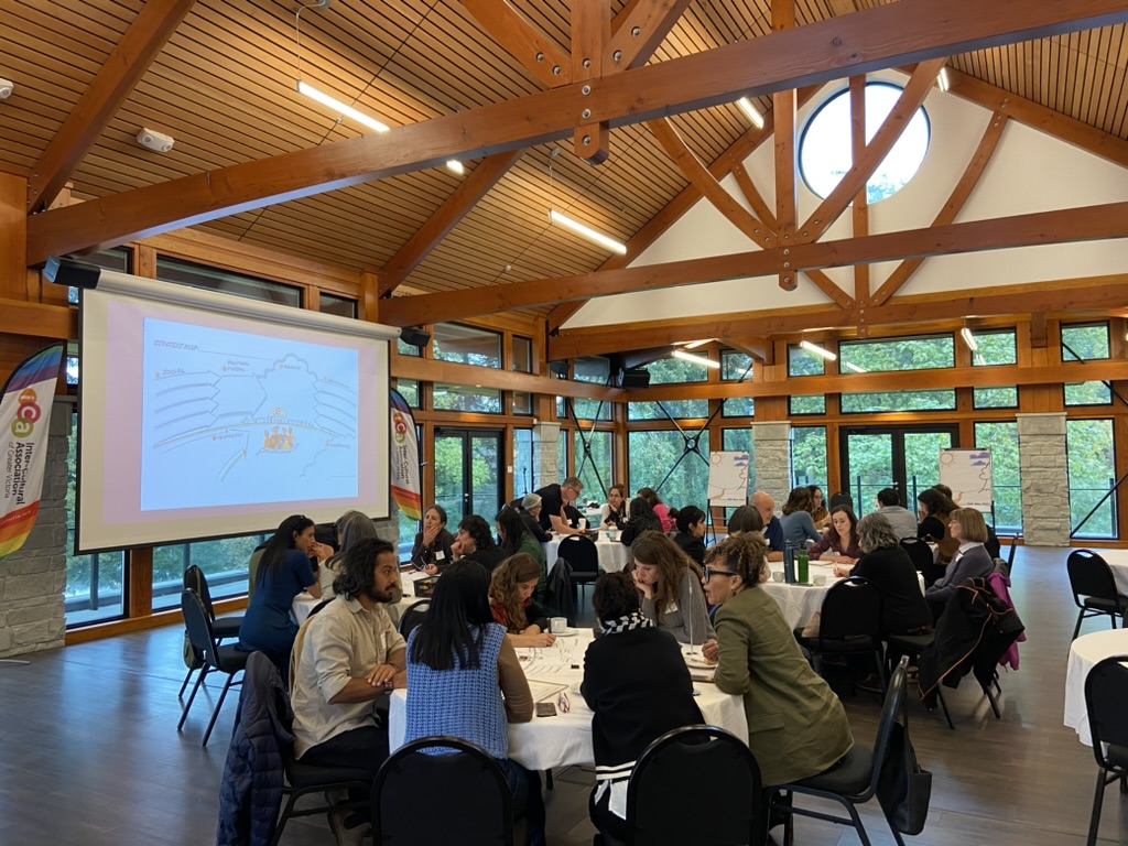In a modern timberframe conference hall, small groups of professionals are in discussion over round tables.