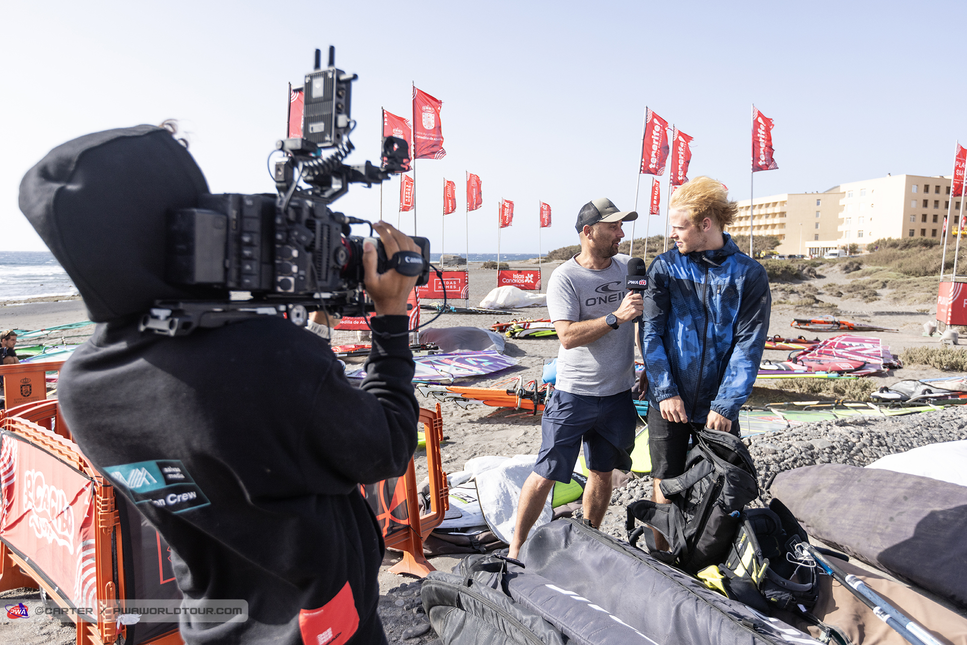 Cameraman filming an interviewer holding a microphone while talking to a windsurfer on a beach with windsurfing gear and red flags in the background.