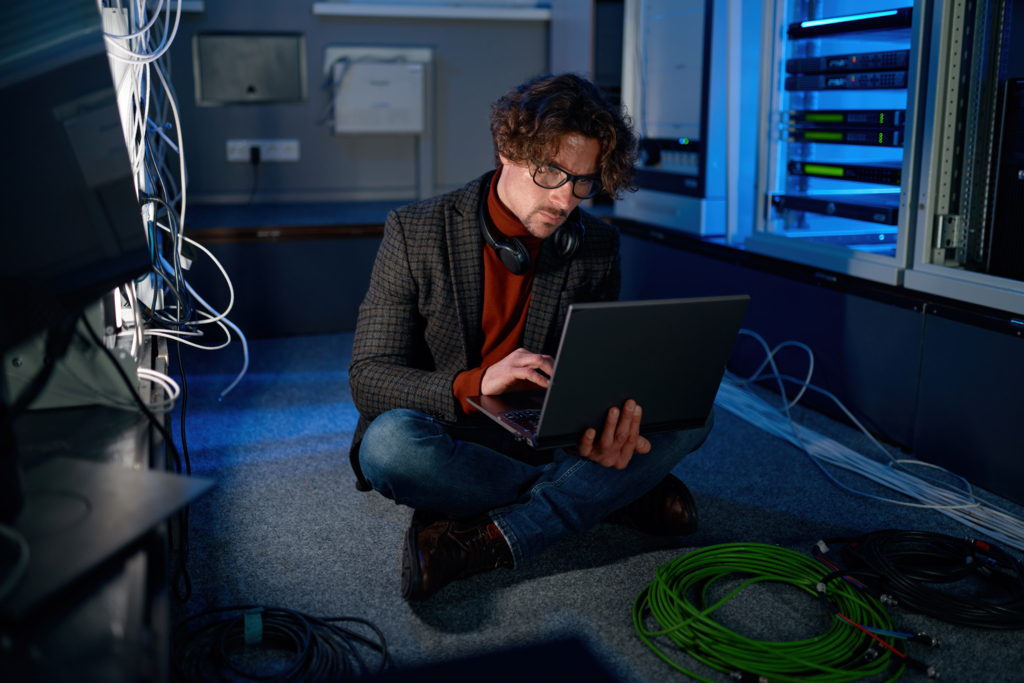 Ein IT-Administrator mit Brille und Kopfhörern sitzt im Schneidersitz auf dem Boden eines Rechenzentrums. Er arbeitet konzentriert an einem Laptop, während um ihn herum Kabelrollen liegen und im Hintergrund beleuchtete Server-Racks zu sehen sind.