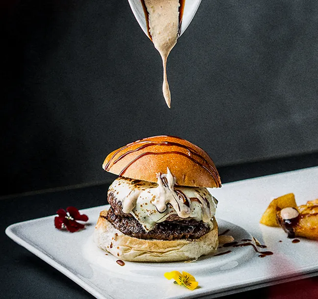 Hamburguesa gourmet con queso derretido, salsa y flores comestibles en un plato blanco.
