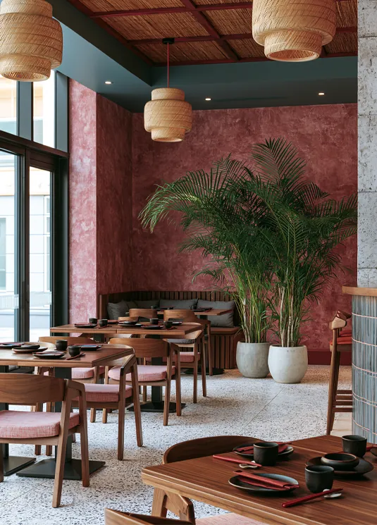 Interior de restaurante con mesas de madera, sillas, grandes plantas y lámparas colgantes de mimbre.