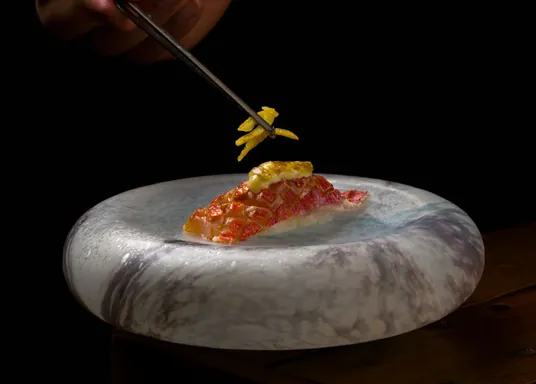 Una mano con palillos sostiene un aderezo sobre pescado servido en un plato de mármol.