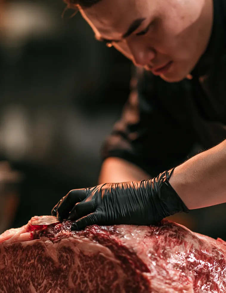 Cocinero con guantes negros manipulando un trozo grueso de carne con marmoleo.
