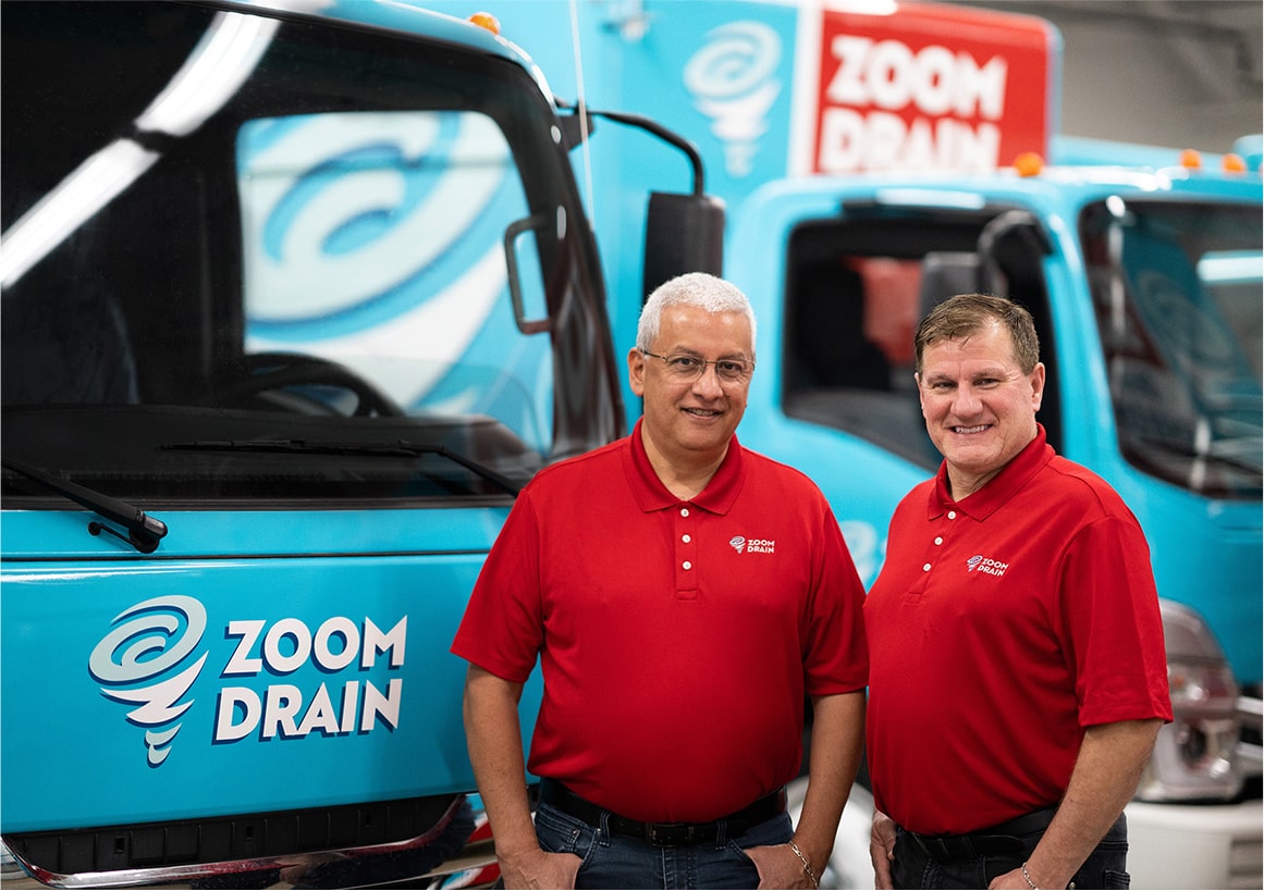 Two male employees in red shirts standing beside Zoom Drain service trucks.