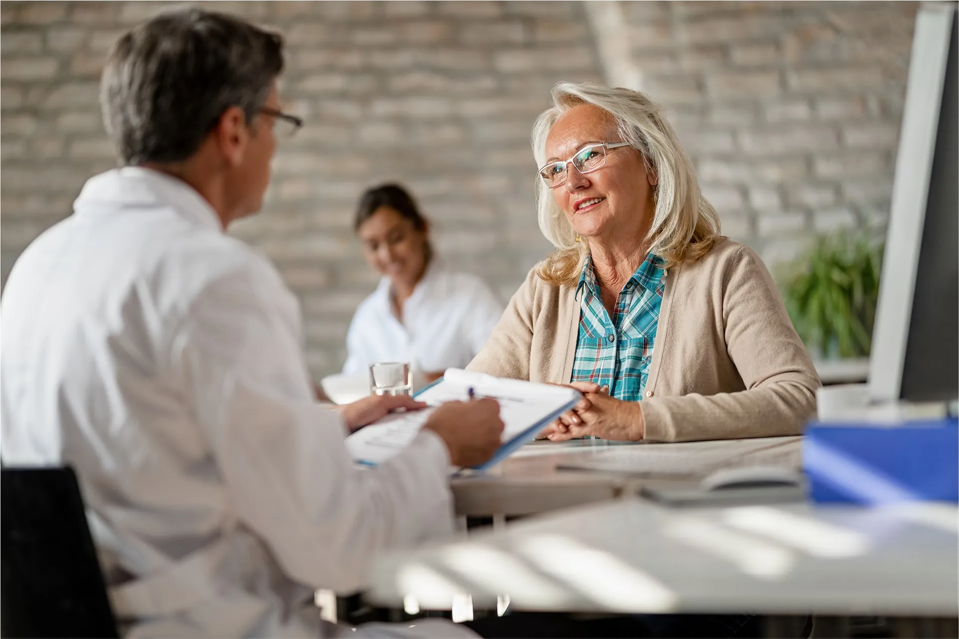 Senior woman having a consultation with a male doctor holding a clipboard, while another medical professional is in the background.