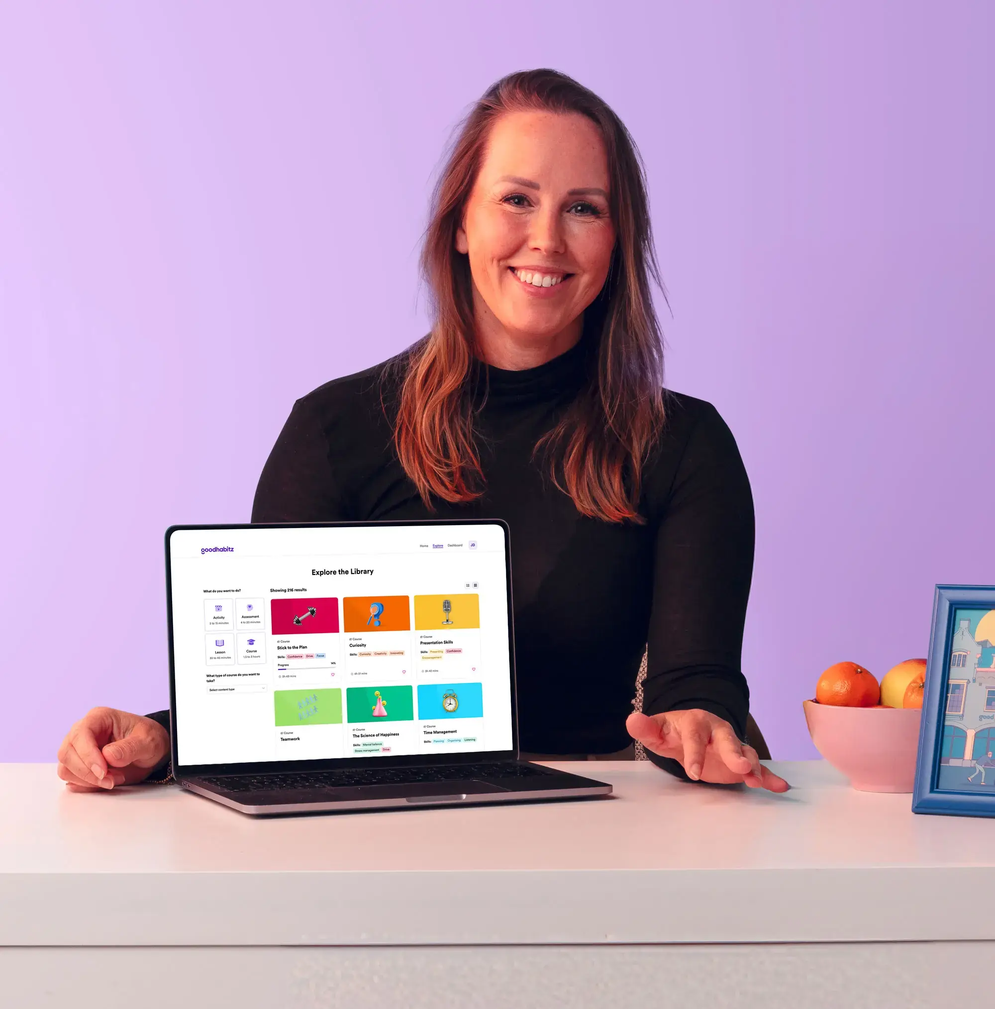 Person sitting at a desk with a laptop displaying the GoodHabitz online learning library interface. The person’s face is intentionally blurred for privacy. On the desk are a white lamp, a bowl of fruit, and a framed picture, all against a pastel purple background.
