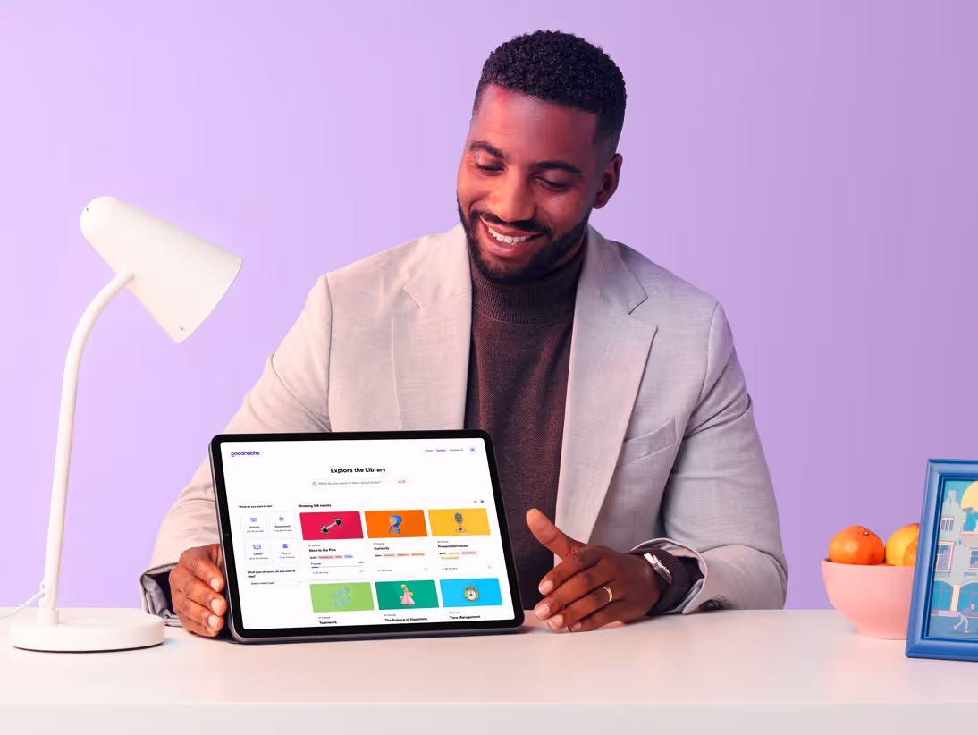 Smiling man in a light blazer sitting at a desk with a tablet showing the GoodHabitz digital learning platform interface, surrounded by a desk lamp, a bowl of fruit, and a framed illustration, with a soft purple background.