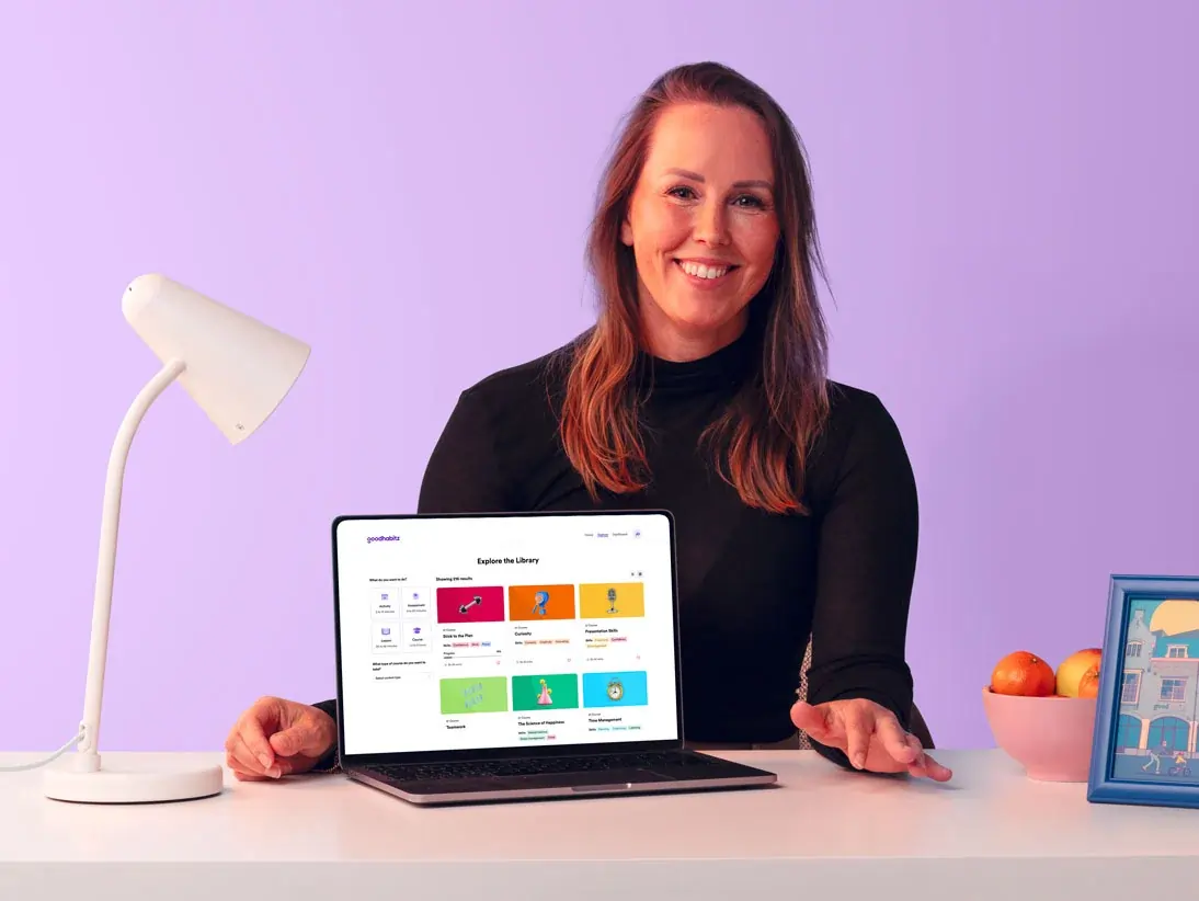 Person sitting at a desk with a laptop displaying the GoodHabitz online learning library interface. The person’s face is intentionally blurred for privacy. On the desk are a white lamp, a bowl of fruit, and a framed picture, all against a pastel purple background.