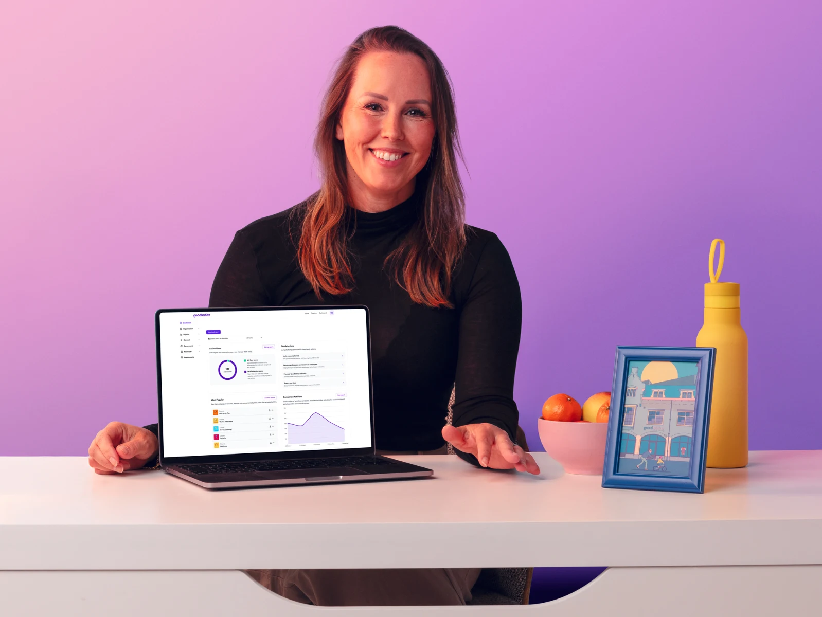 Friendly woman sitting at a desk showing the GoodHabitz dashboard on her laptop.