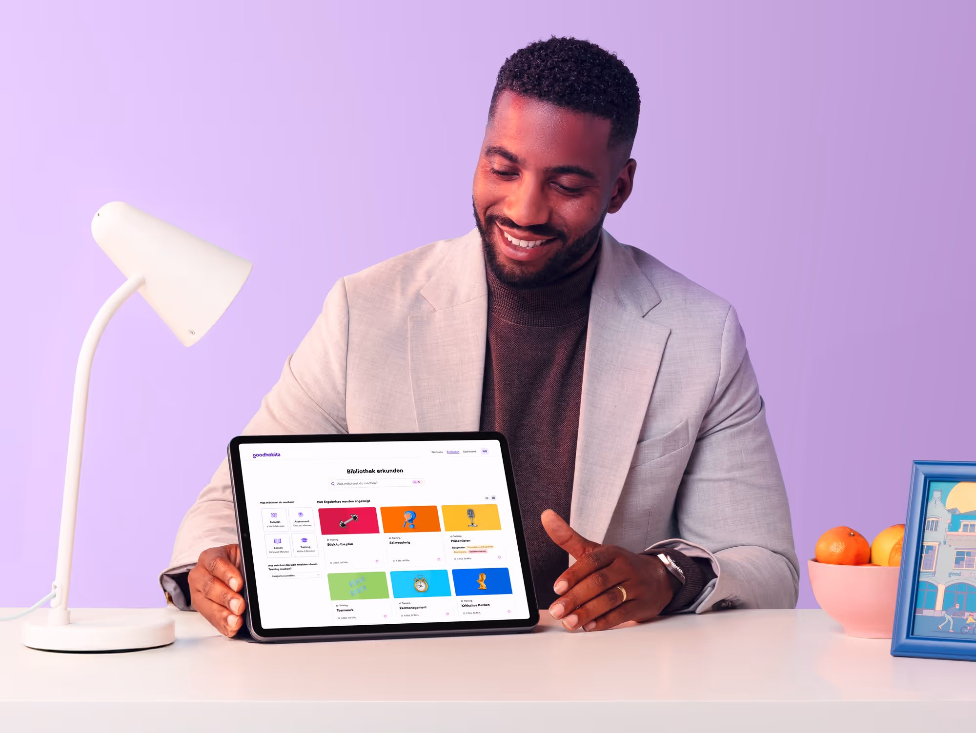 Smiling man in a light blazer sitting at a desk with a tablet showing the GoodHabitz digital learning platform interface, surrounded by a desk lamp, a bowl of fruit, and a framed illustration, with a soft purple background.