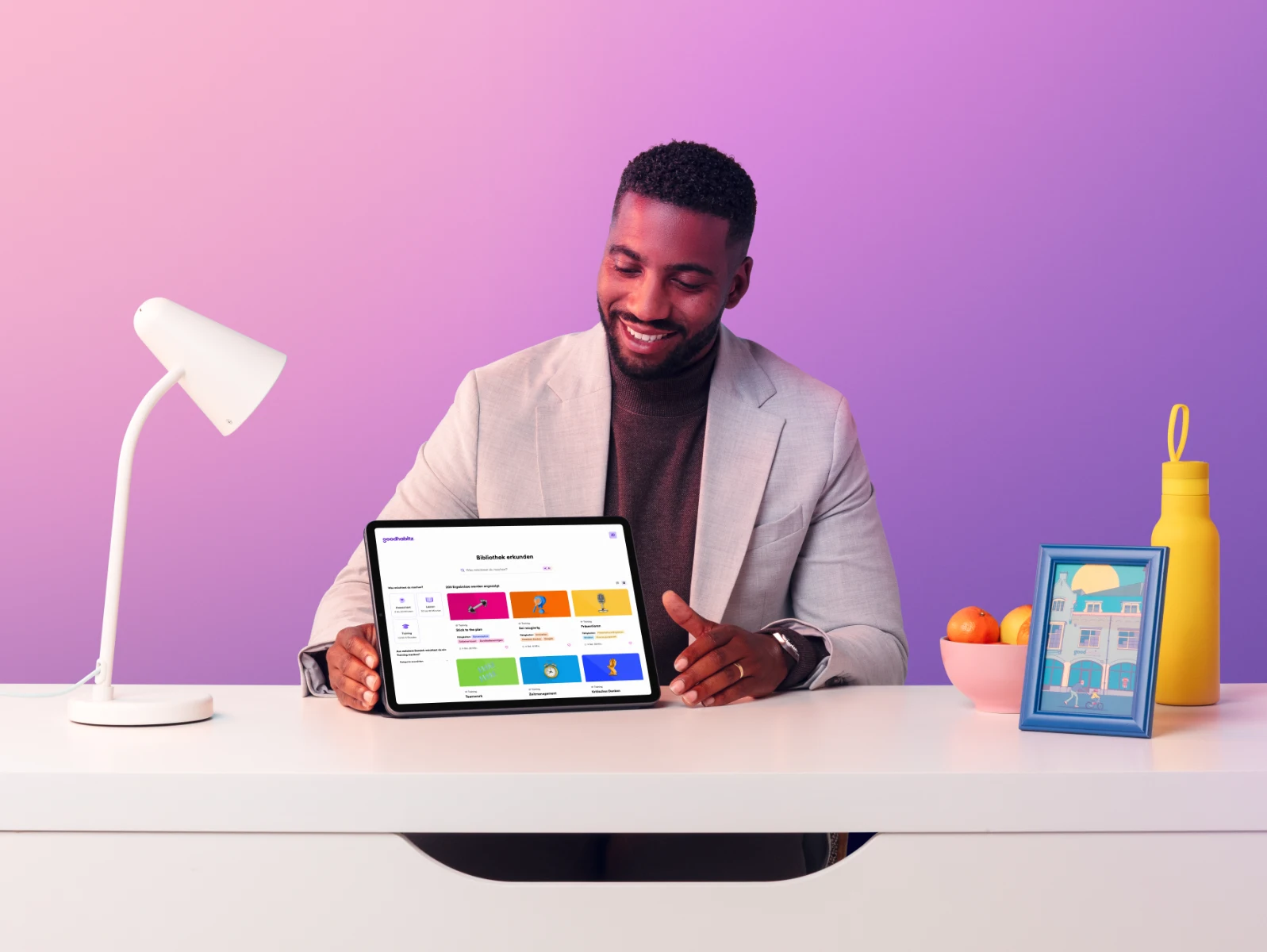 Smiling man in a light blazer sitting at a desk with a tablet showing the GoodHabitz digital learning platform interface, surrounded by a desk lamp, a bowl of fruit, and a framed illustration, with a soft purple background.