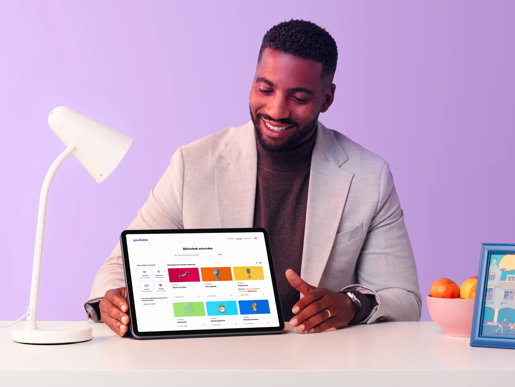 Smiling man in a light blazer sitting at a desk with a tablet showing the GoodHabitz digital learning platform interface, surrounded by a desk lamp, a bowl of fruit, and a framed illustration, with a soft purple background.