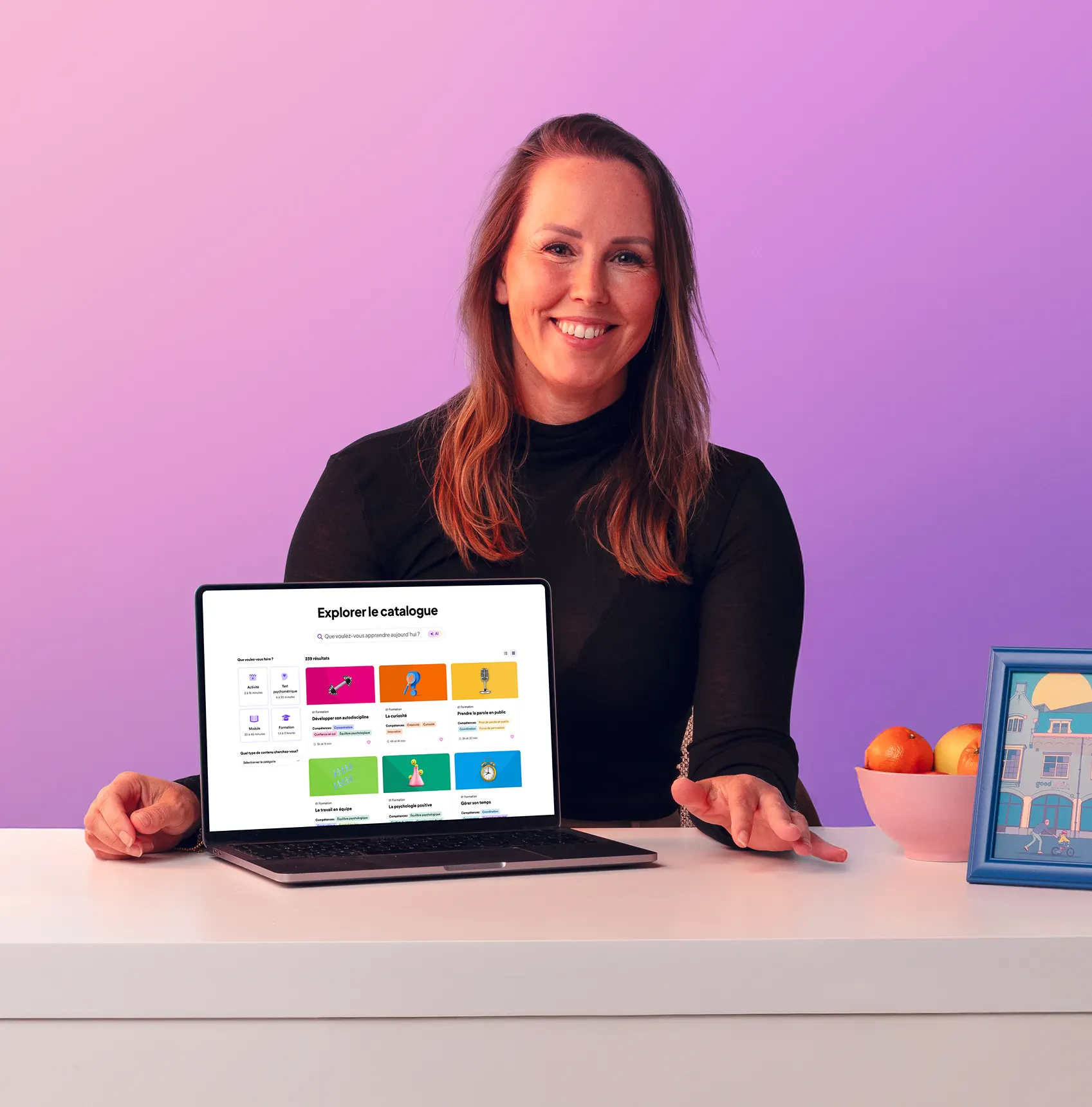 Personne assise à un bureau avec un ordinateur portable affichant l'interface de la bibliothèque d'apprentissage en ligne GoodHabitz. Le visage de la personne est volontairement flouté pour préserver la confidentialité. Sur le bureau, une lampe blanche, un bol de fruits et une photo encadrée, le tout sur un fond violet pastel.