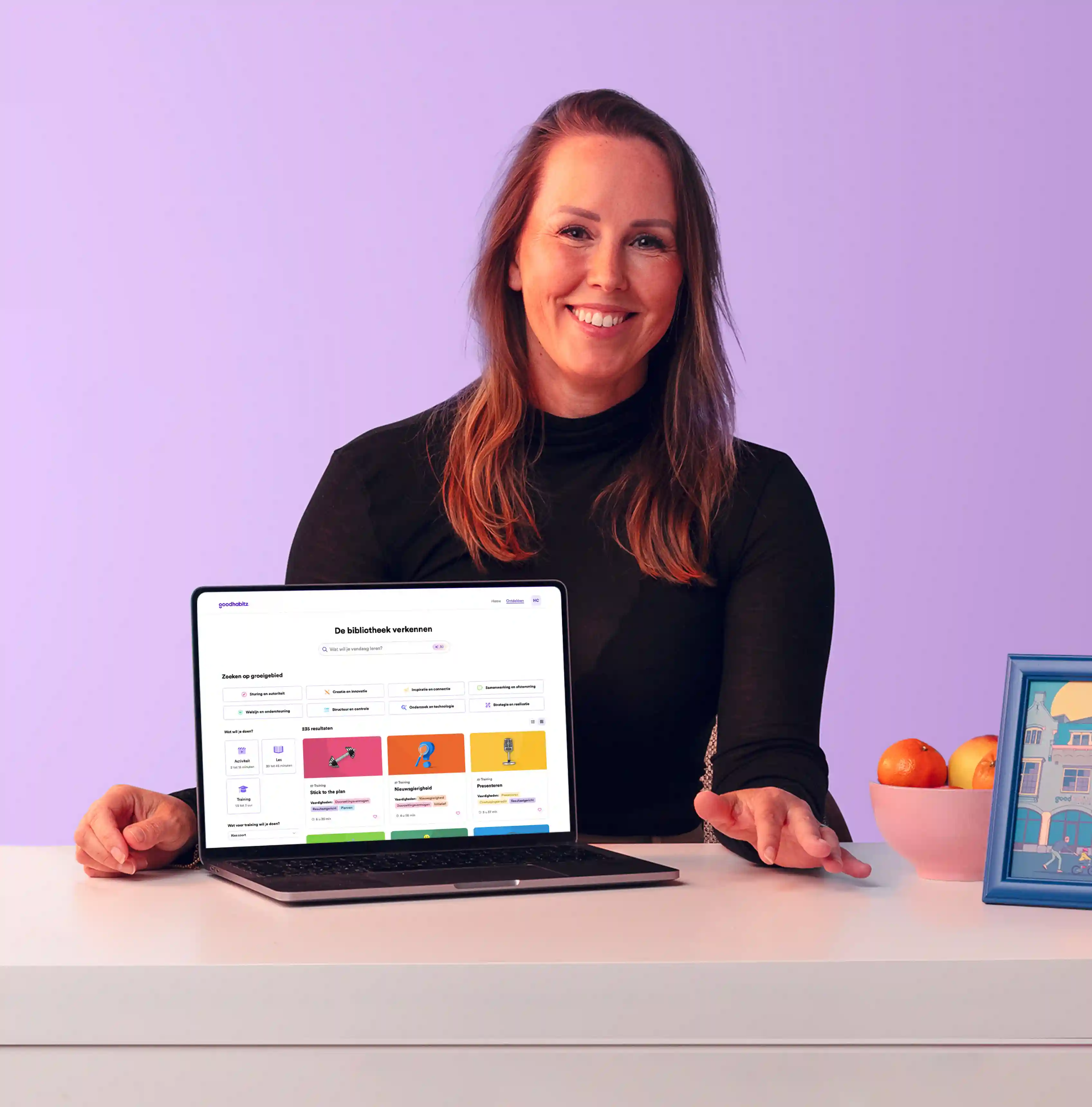 Persoon zit aan een bureau met een laptop die de interface van de GoodHabitz online le bibliotheek toont. Het gezicht van de persoon is bewust vervaagd voor privacy. Op het bureau staan een witte lamp, een schaal fruit en een ingelijste foto, tegen een pastelpaarse achtergrond.