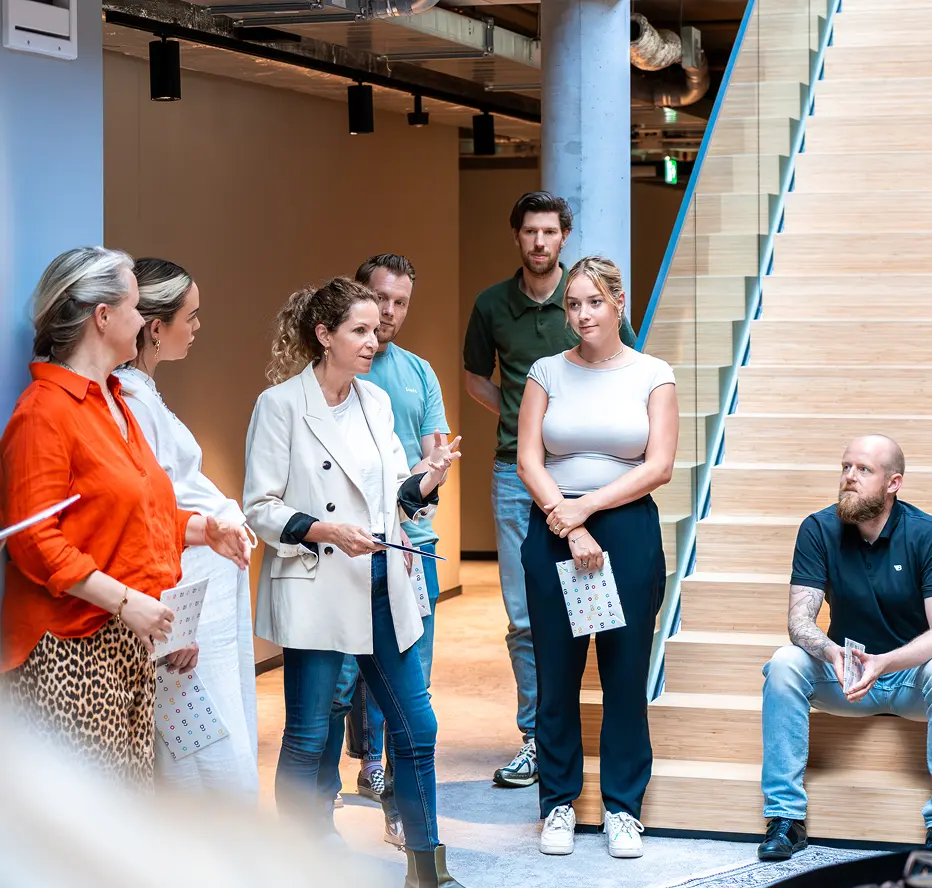 Group of six people standing and sitting near wooden stairs in a modern office, engaged in a discussion.