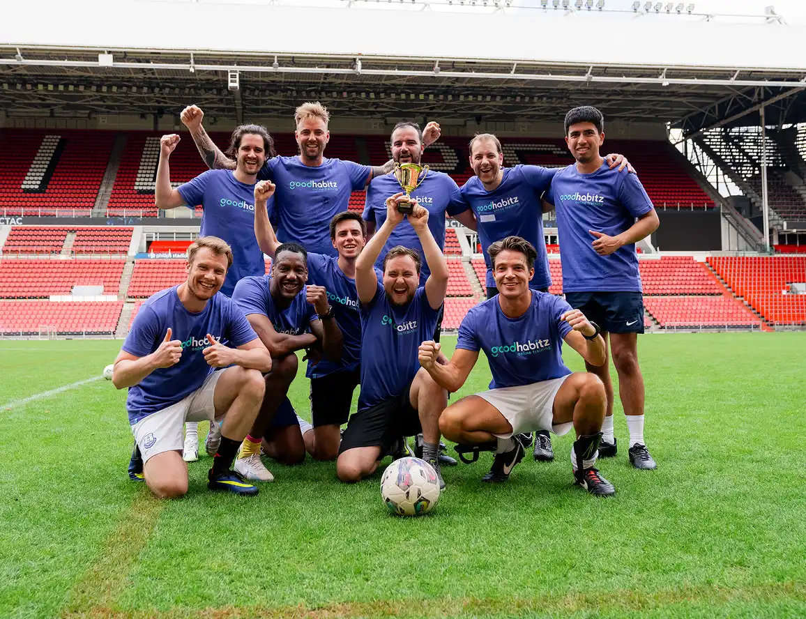 A group of ten men in blue 'goodhabitz' shirts celebrate on a soccer field, holding a trophy and posing with a soccer ball in front of stadium seats.