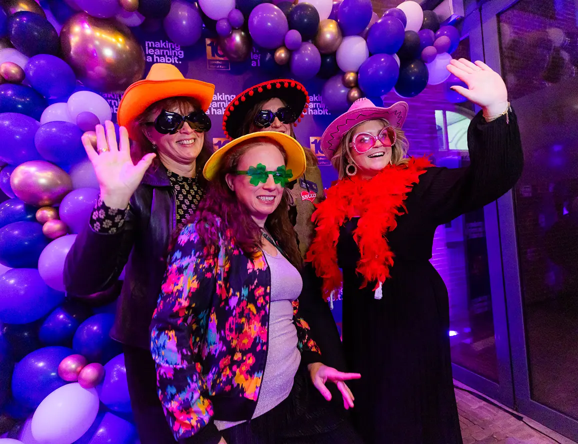 Four women wearing colorful hats, oversized glasses, and a feather boa posing and waving in front of a purple and gold balloon arch.