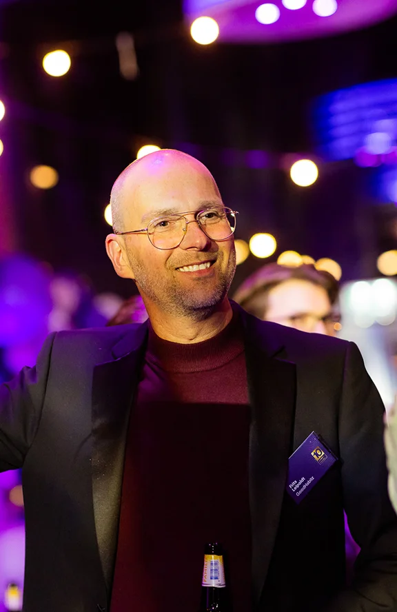 Smiling man wearing glasses, a dark blazer, and a maroon shirt at an indoor event with blurred lights in the background.