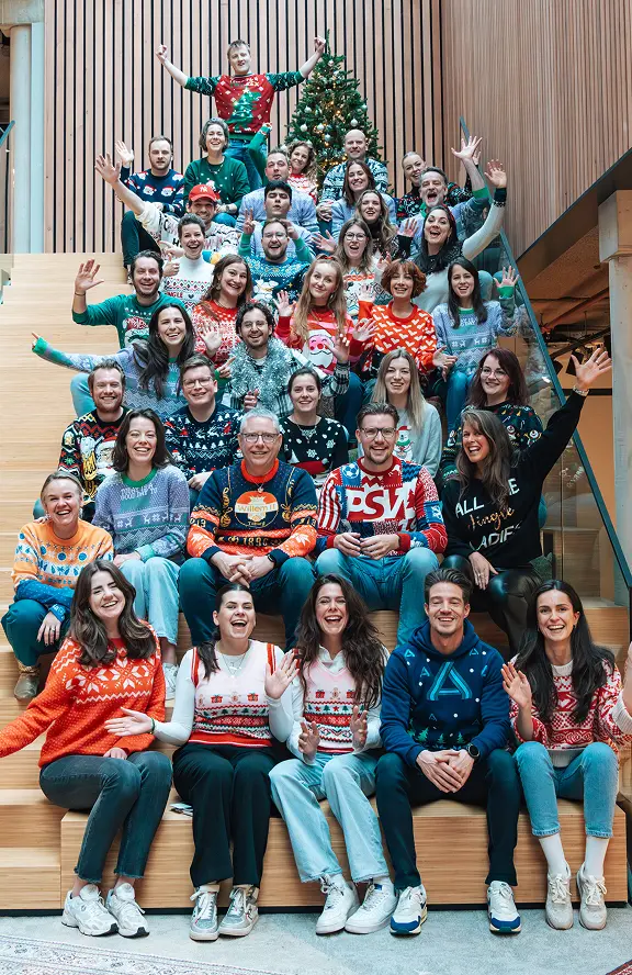 Large group of smiling people wearing colorful Christmas sweaters, seated on wooden stairs in front of a decorated Christmas tree, waving at the camera.