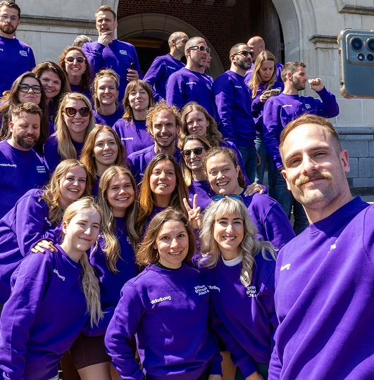 Group of smiling people in matching purple sweatshirts taking a selfie outdoors in front of a stone building.