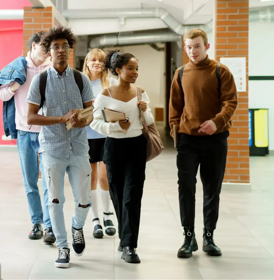 A group of diverse young students walking and talking together down a bright school corridor, carrying notebooks and folders.
