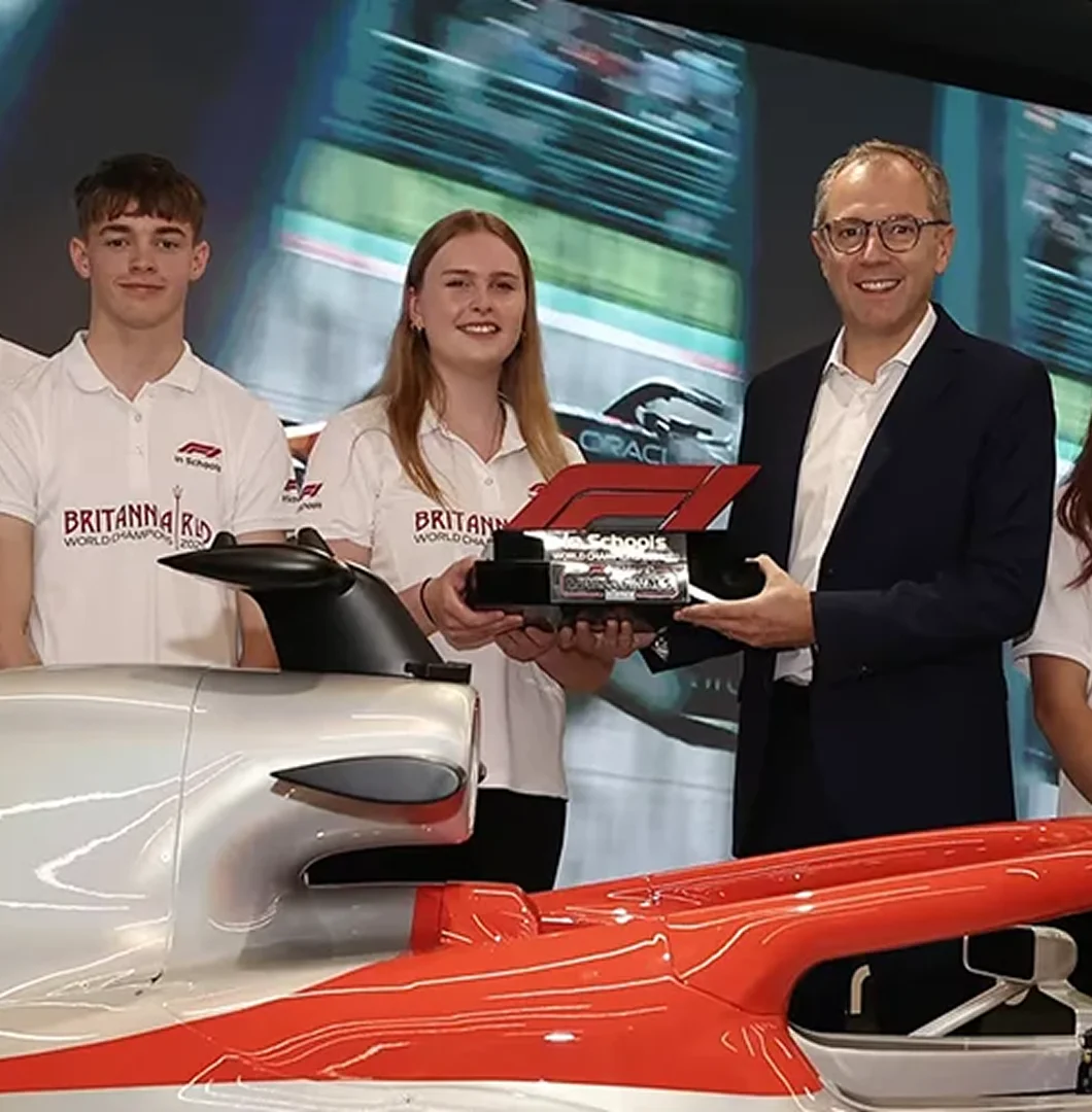 Two young students in white team polo shirts and a smiling man in a suit jacket holding a large red and black F1 in Schools trophy together behind a model racing car.