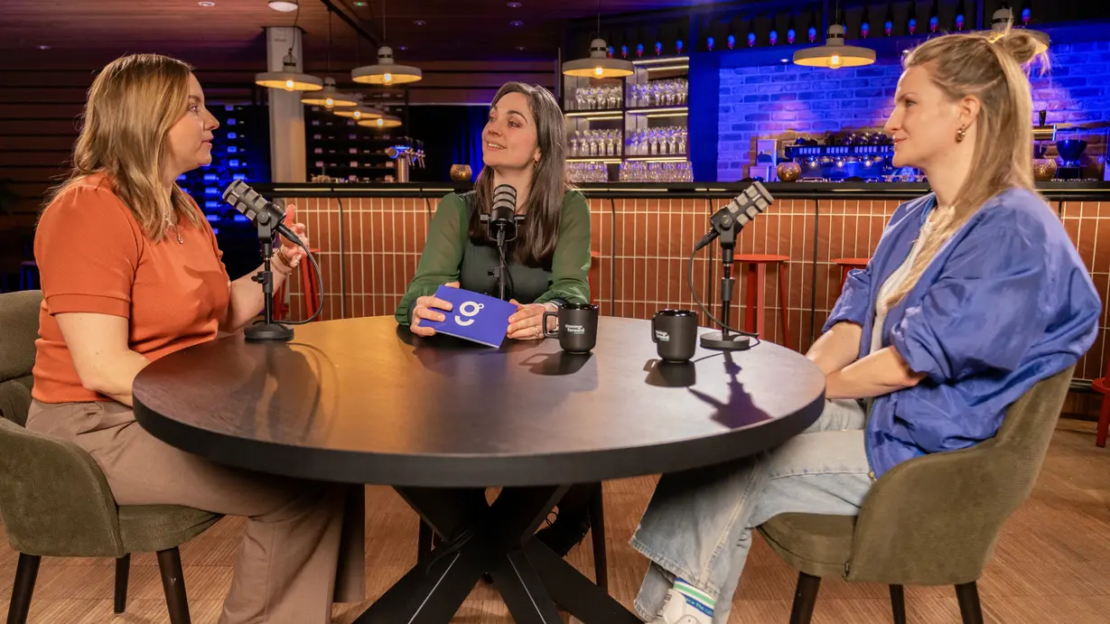 Three people seated around a round black table in a contemporary bar or café, each with a microphone and black mugs in front of them. The scene suggests a relaxed and engaging podcast conversation.