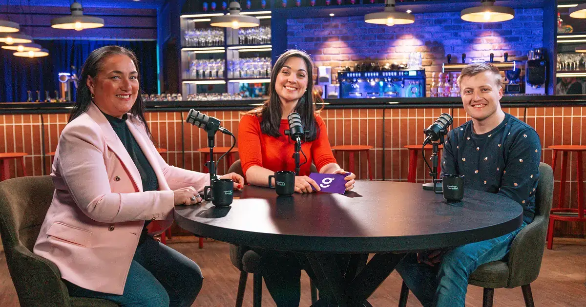 Three people sit around a round black table in a modern bar or café setting equipped for podcast recording. Each person has a microphone and a black mug in front of them