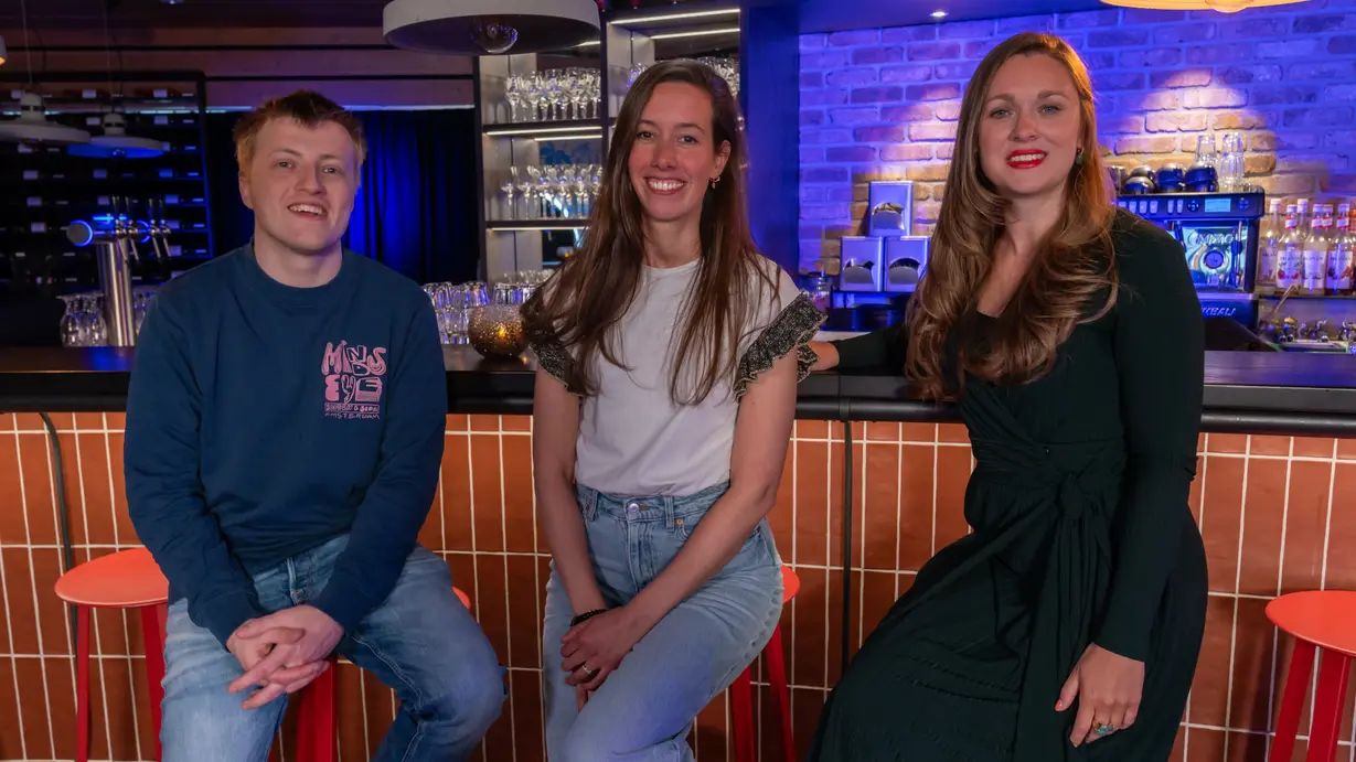Three people sit on stools at a modern café or bar counter, posing for a photo. The person on the right, with long red hair and wearing a black dress, is behaviour change expert Amy-Jane Gielen.