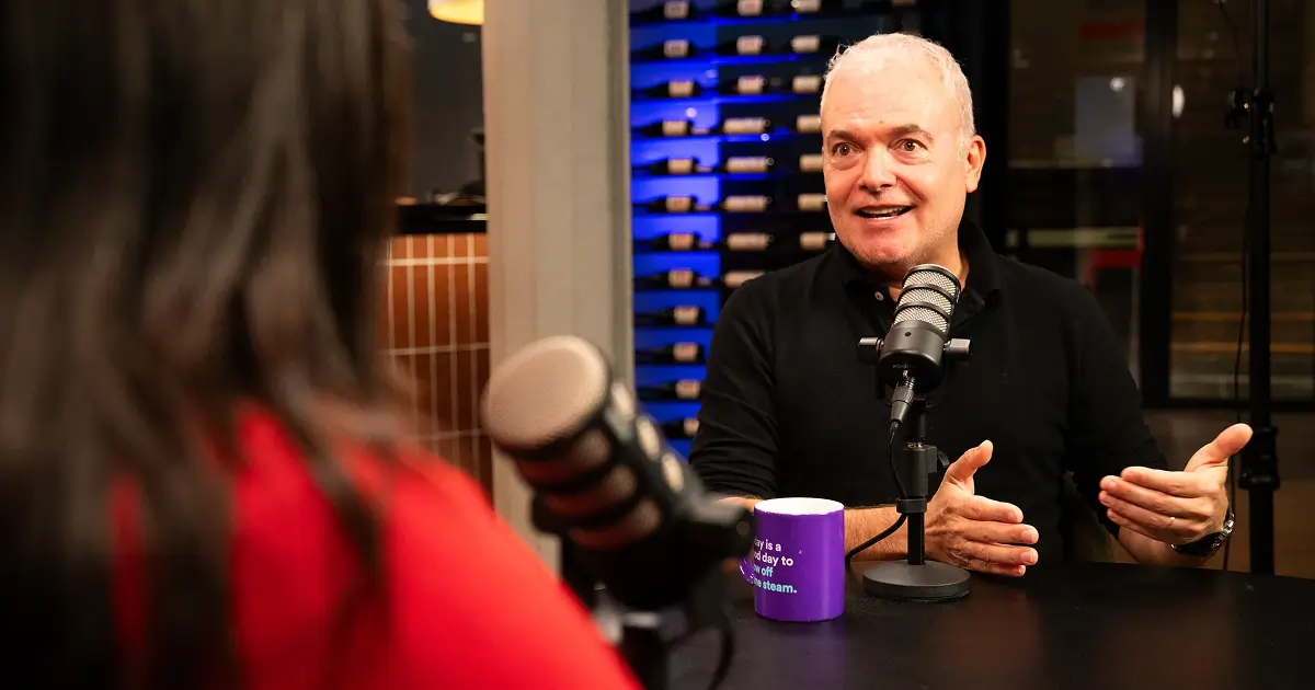Podcast episode screenshot showing Marteen Franken in conversation at a round table, both speaking into microphones. A purple mug is visible on the table, set in a warmly lit recording studio with a backdrop of shelves and modern decor.