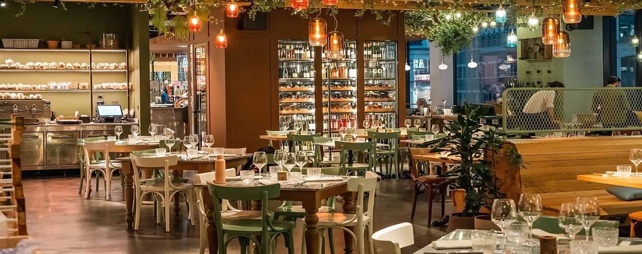 Un restaurant élégant avec des tables en bois, des chaises vertes et blanches, et des verres à vin disposés. L'éclairage est chaleureux avec des lampes suspendues et des plantes décoratives. Une étagère de bouteilles de vin est visible à l'arrière-plan.