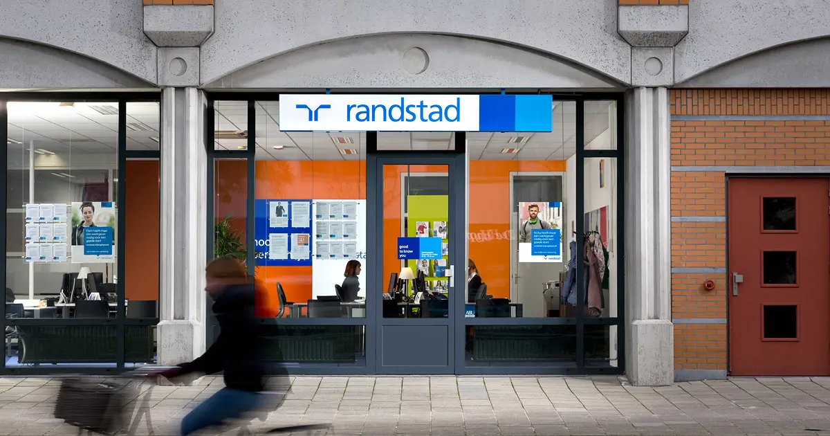 Façade d'une agence Randstad avec de grandes fenêtres vitrées. À l'intérieur, on aperçoit des bureaux et des affiches sur les murs. Une personne passe à vélo devant l'agence.