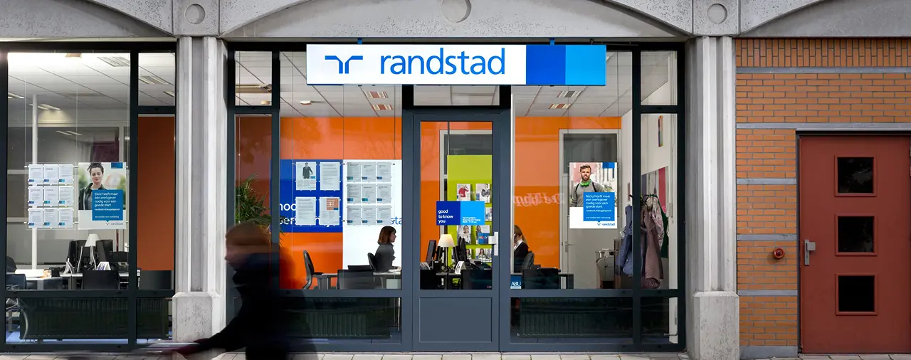Façade d'une agence Randstad avec de grandes fenêtres vitrées. À l'intérieur, on aperçoit des bureaux et des affiches sur les murs. Une personne passe à vélo devant l'agence.