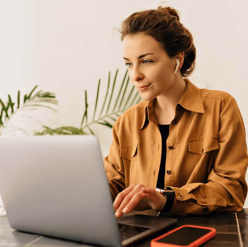 Frau mit brauner Bluse am Laptop, drahtlose Kopfhörer, Smartphone auf Tisch, grüne Pflanze im Hintergrund.
