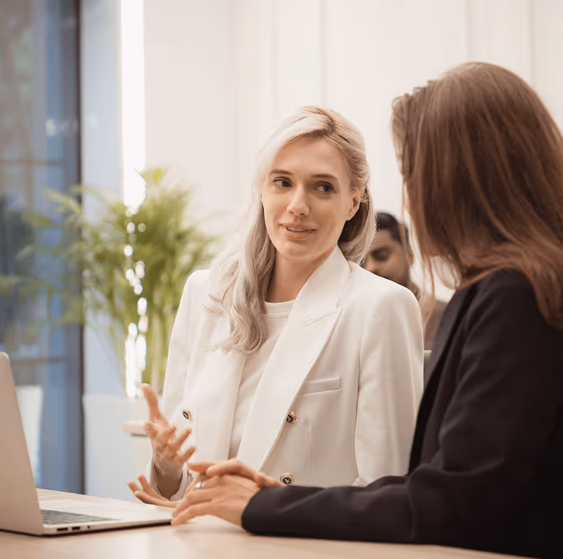 Zwei Frauen im Gespräch, eine in weißem Blazer erklärt, während andere zuhört; Pflanzen im Hintergrund, Laptop auf Tisch.