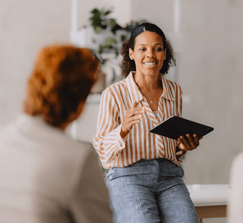 Frau in gestreifter Bluse spricht lächelnd und gestikuliert mit Tablet in der Hand, während sie vor einem unscharfen Kollegen sitzt.