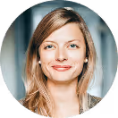 Lächelnde Frau mit blonden Haaren und Perlenohrringen, vor verschwommenem Hintergrund.