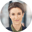 Lächelnde Person mit kurzem Haar vor verschwommenem Hintergrund in Geschäftskleidung.