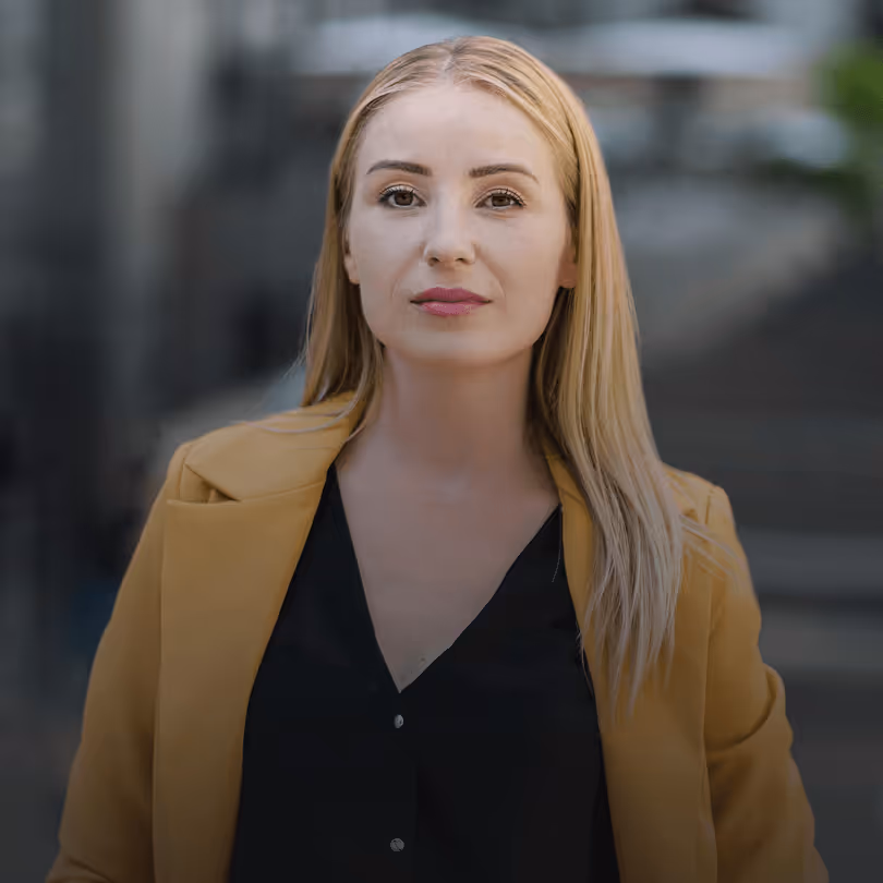 Frau mit langen blonden Haaren in gelbem Blazer steht selbstbewusst draußen vor unscharfem Hintergrund.